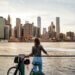 Individual standing by a railing with a bicycle, gazing at the New York City skyline across the water.